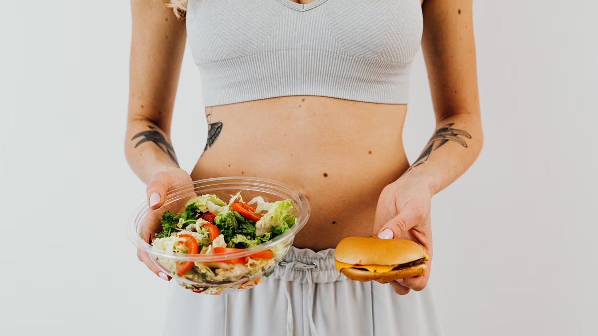 A woman in a sports bra holding a salad and cheeseburger, representing healthy vs junk food choices.